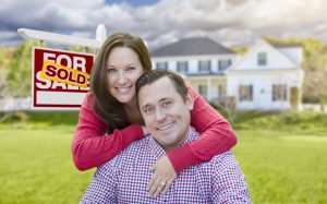 Happy Couple In Front Of Sold For Sale Real Estate Sign And Beautiful House.